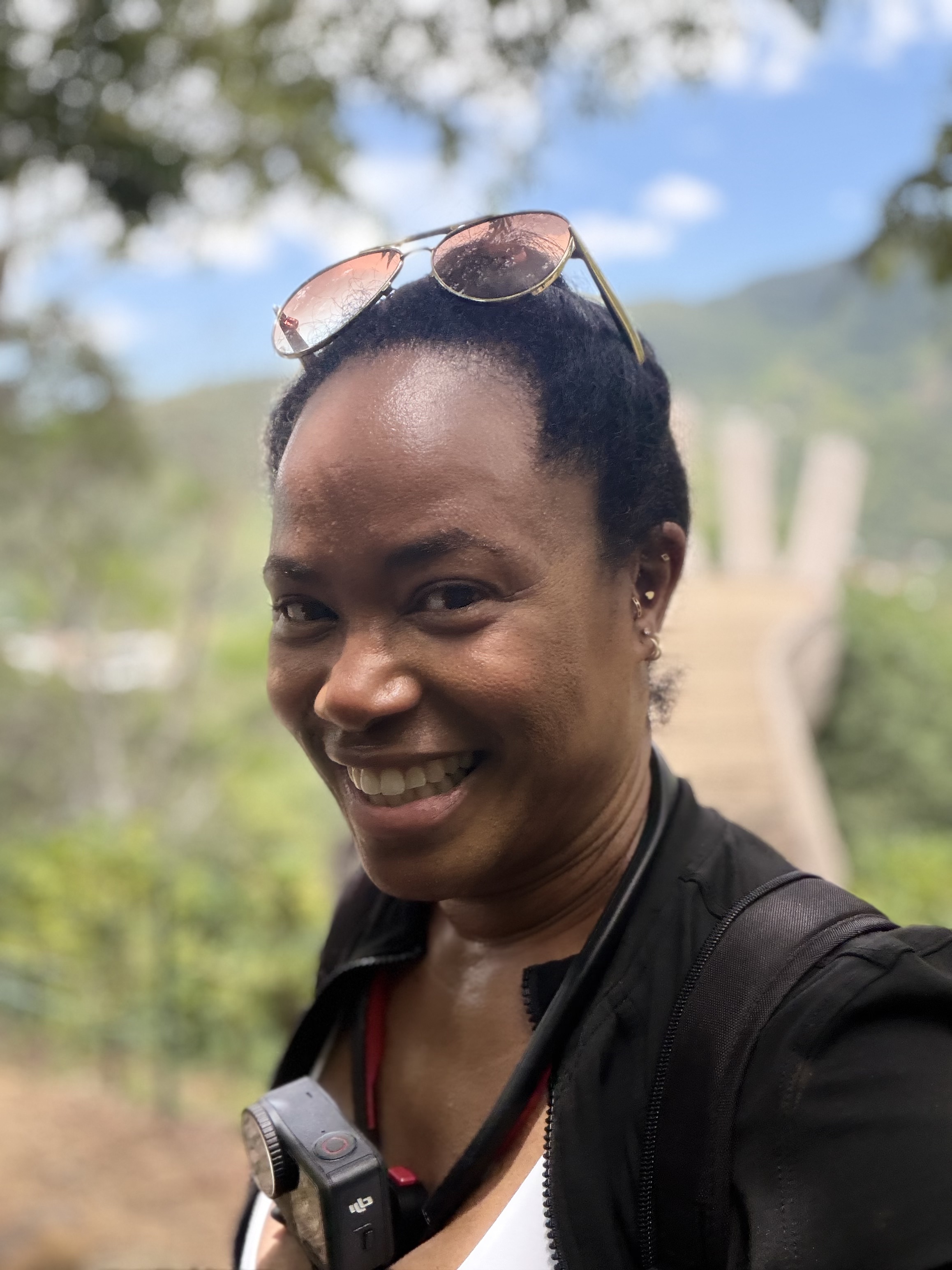 Electa smiling outdoors during a travel moment in a scenic, green environment.